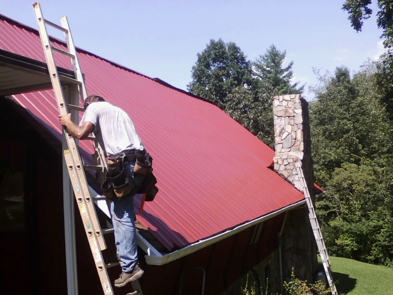 Roofer on a ladder beside a red metal roof and stone chimney — Soffit & Fascia Repair by Roofing Brothers in Mount Carmel